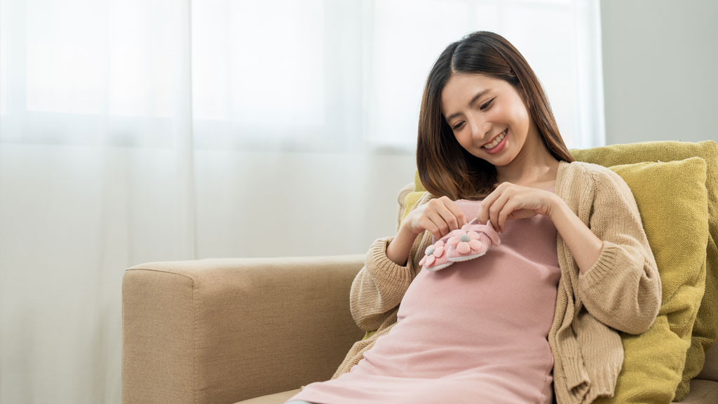 Pregnant lady sitting on a sofa holding baby shoes over her tummy