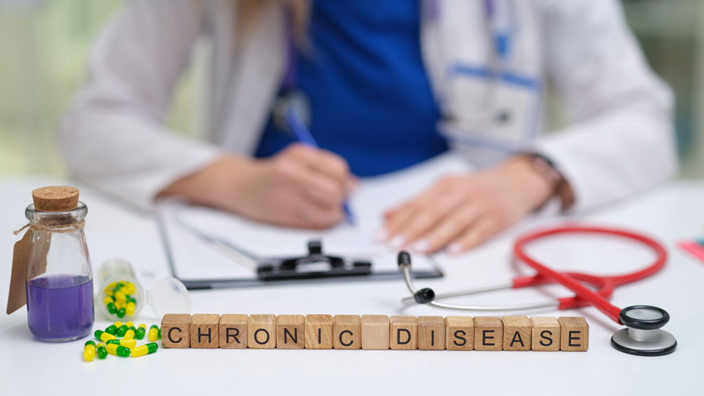 Doctor sitting at a desk making notes with Chronic Disease spelled out on wood blocks